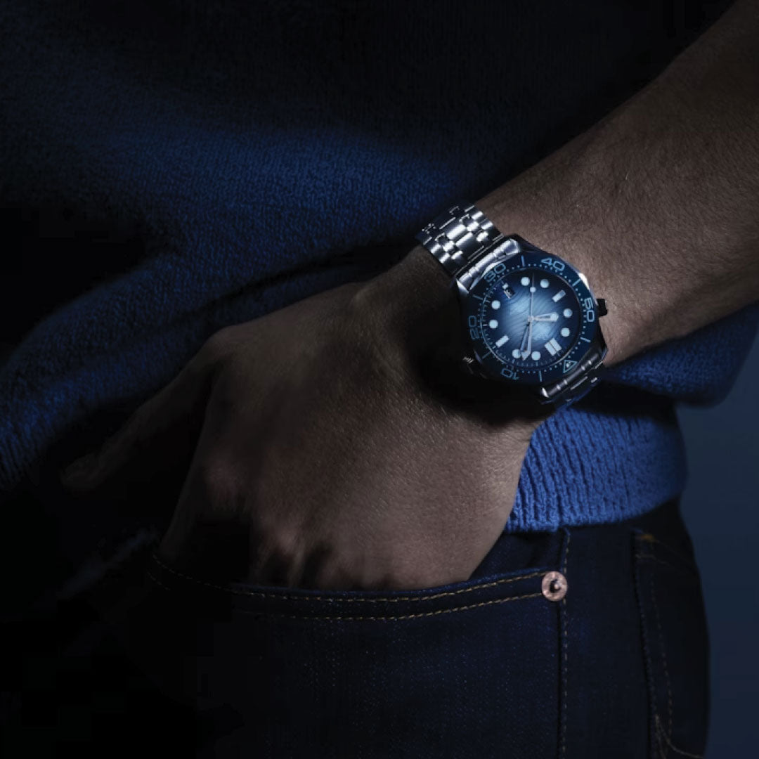 In a dimly lit setting, an individual wearing a blue sweater and jeans has their right hand in a pocket, elegantly presenting the OMEGA Seamaster Diver 300M Summer Blue Co-Axial Master Chronometer 42mm Watch.