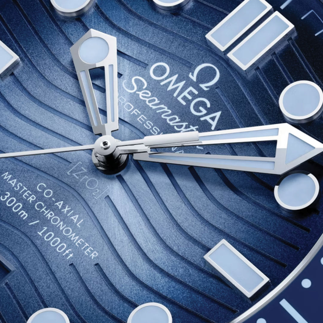 The OMEGA Seamaster Diver 300M Summer Blue Co-Axial Master Chronometer 42mm watch features a detailed close-up of its captivating wave-patterned dial. It proudly displays the "Omega Seamaster Professional" text, alongside luminescent hour markers and hands, and specifies a depth rating of "300m/1000ft.