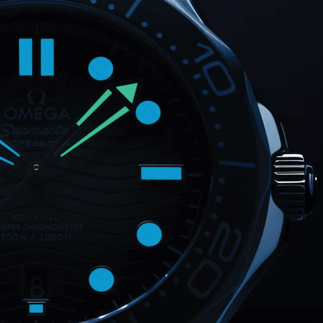 The OMEGA Seamaster Diver 300M Summer Blue Co-Axial Master Chronometer 42mm Watch is shown in a close-up view, with its face glowing in the dark. The luminescent hour markers and hands shine in Summer Blue and green, while the Co-Axial Master Chronometer calibre 8800 branding and some numerals are subtly visible in the low light.