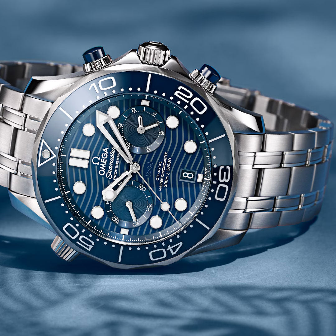 A close-up of the OMEGA Seamaster Diver 300M Co-Axial Master Chronometer Chronograph 44mm watch, featuring a blue wave-patterned dial, silver indices, and a stainless steel bracelet on a light blue surface.