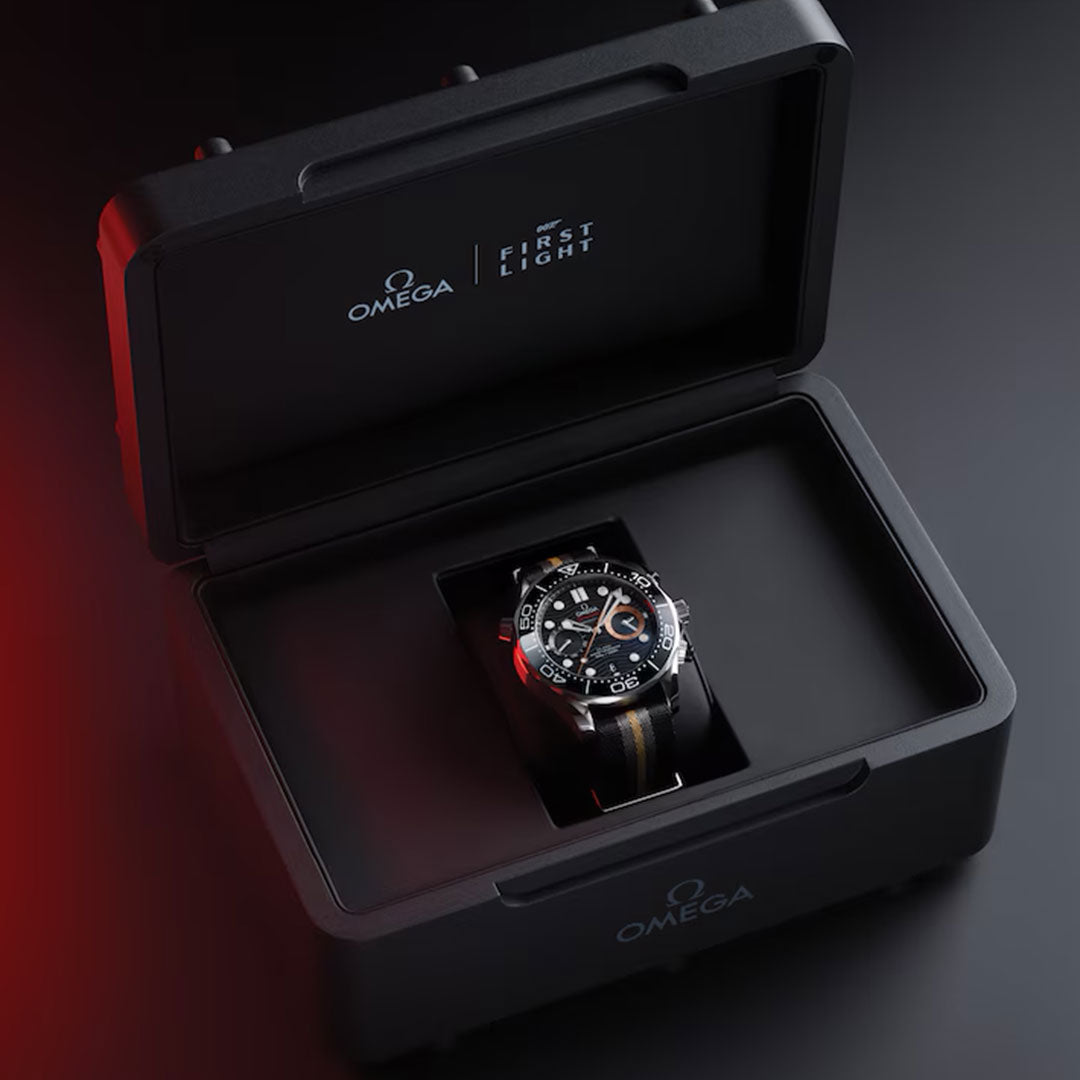 An OMEGA Seamaster Diver 300M 007 First Light Edition Co-Axial Master Chronometer Chronograph 44mm watch, featuring a black strap, is presented in an open black box adorned with “OMEGA” and “FIRST EIGHT” labeling, set against a dark backdrop with a red light glow on the left.