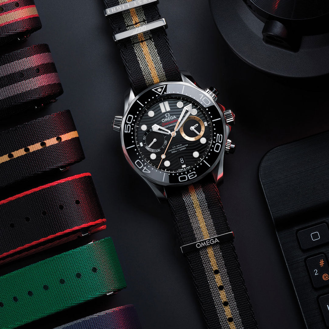 An OMEGA Seamaster Diver 300M 007 First Light Edition Co-Axial Master Chronometer Chronograph 44mm watch with a black striped nylon strap rests beside a laptop keyboard and several colorful striped watch straps on a dark surface.