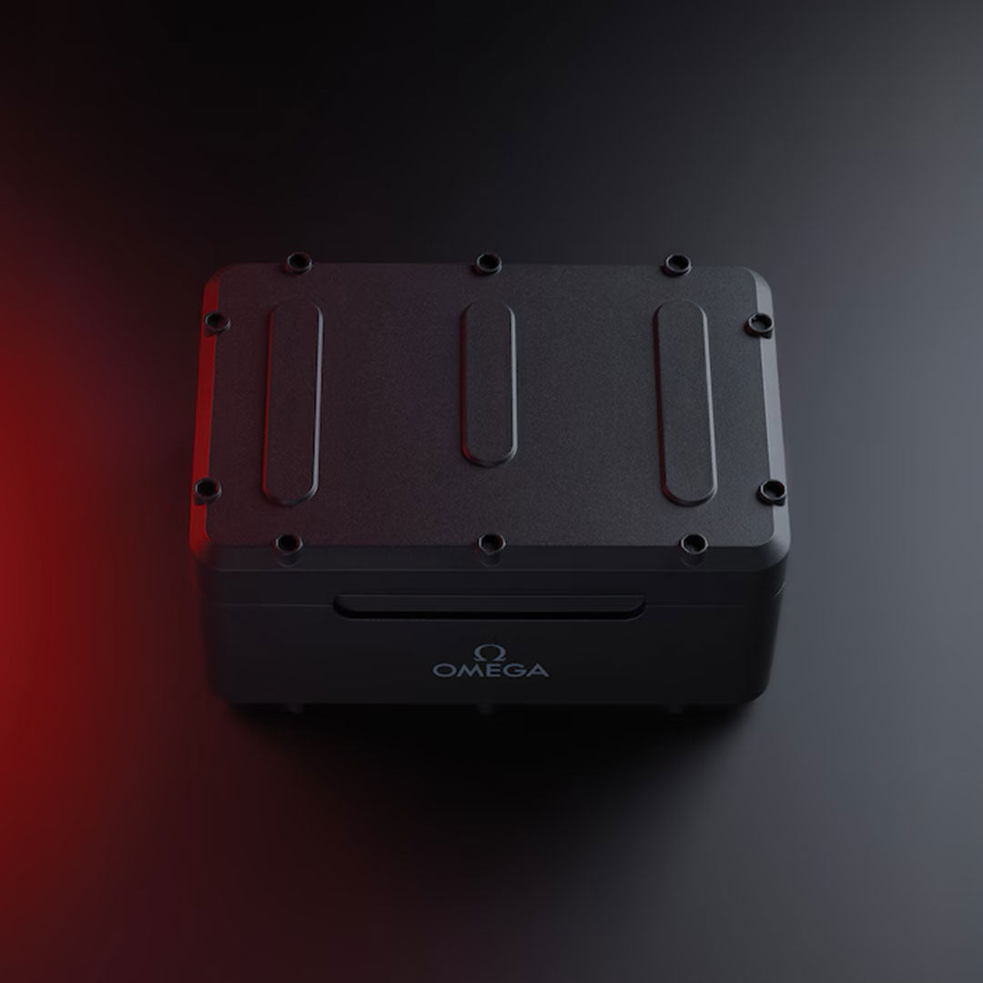 A sleek, black rectangular box labeled “OMEGA” rests on a dark surface with a subtle red light illuminating its left side. Designed in the signature style of the OMEGA Seamaster Diver 300M 007 First Light Edition Co-Axial Master Chronometer Chronograph 44mm watch packaging, the box features rounded edges and prominent screws on its lid.