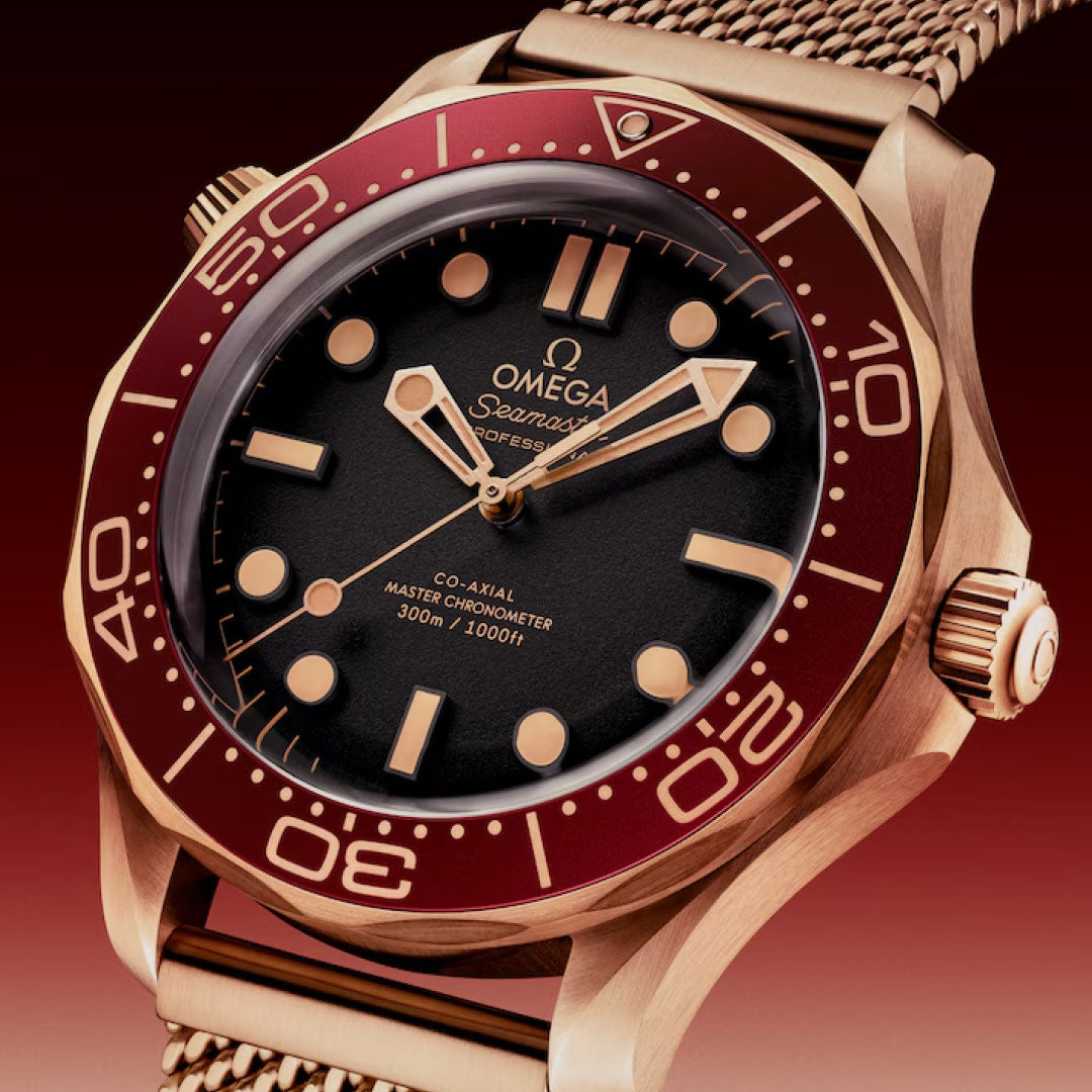 A close-up of the OMEGA Seamaster Diver 300M Co-Axial Master Chronometer 42mm watch shows a red bezel, black dial, gold markers, and a mesh bracelet in Bronze Gold, set against a gradient red background. Luminescent hands and indices complete the look.