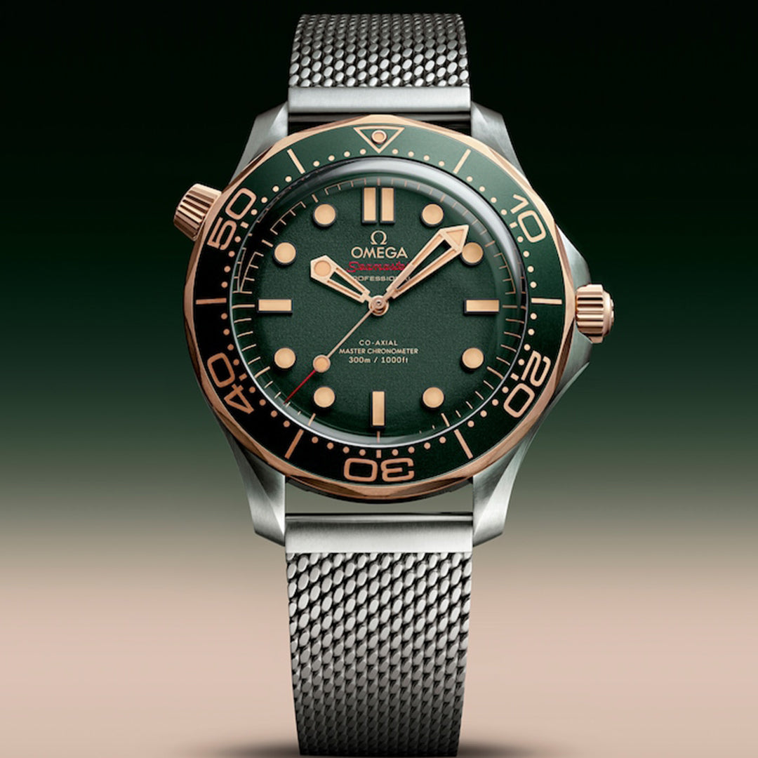 The OMEGA Seamaster Diver 300M Co-Axial Master Chronometer 42mm Watch features a green dial with gold accents on the case, hour markers, bezel, and hands. A silver mesh band complements the design, along with the Seamaster Diver 300M logo for water resistance and depth specifications.