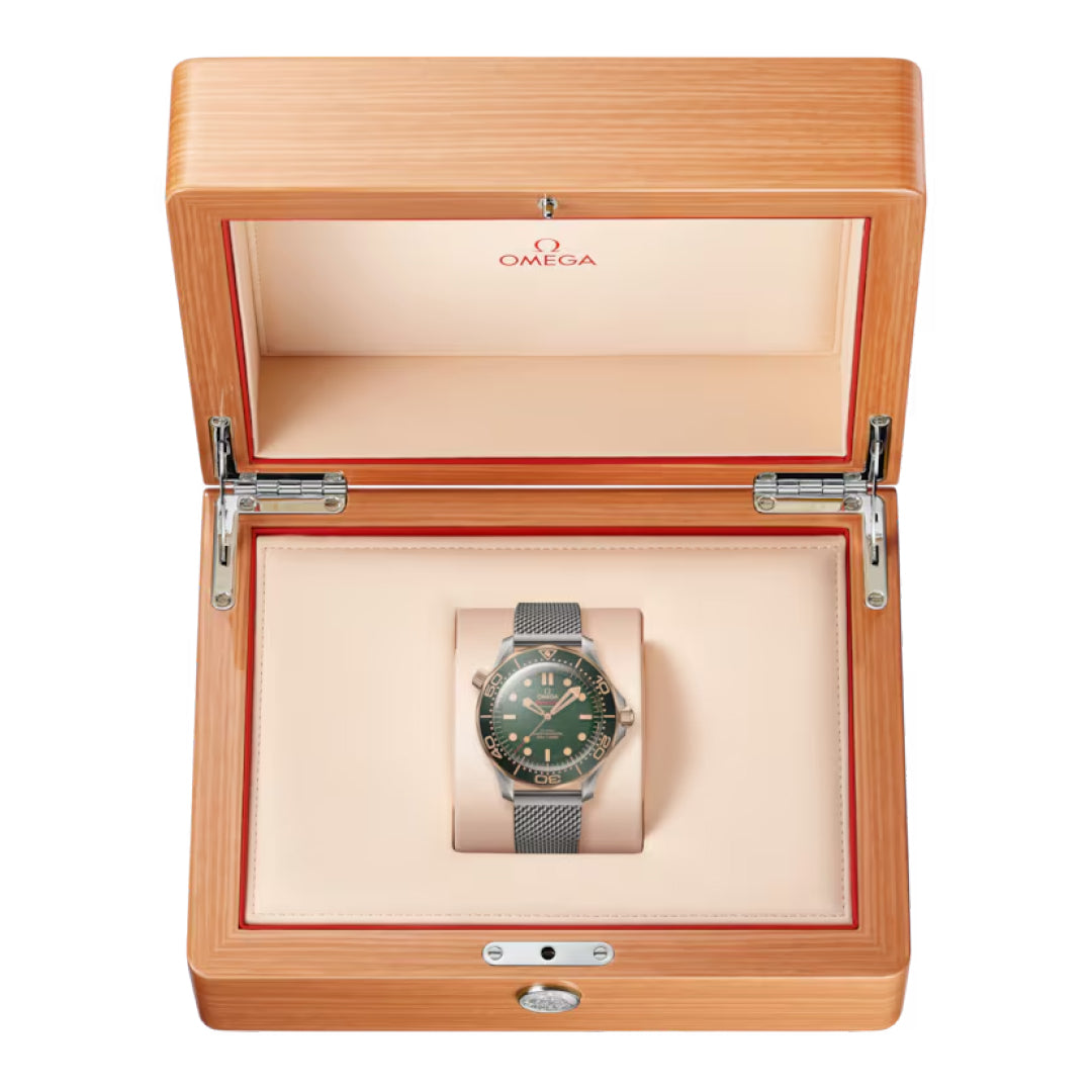The OMEGA Seamaster Diver 300M Co-Axial Master Chronometer 42mm Watch, featuring a green face and metal bracelet, is beautifully displayed in an open wooden Omega box with a light interior. It sits within a padded compartment, ideal for lovers of timeless luxury.