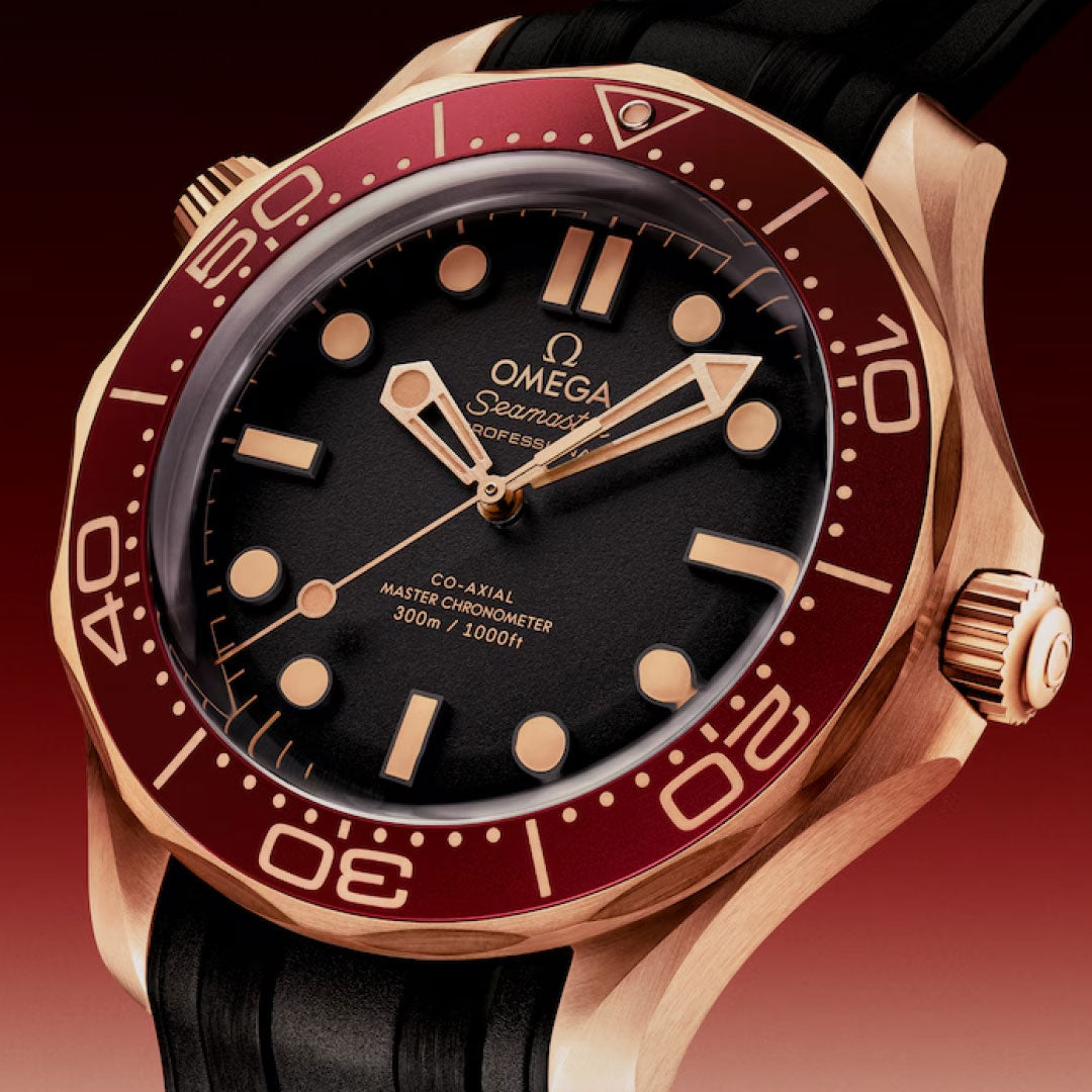 A close-up of the OMEGA Seamaster Diver 300M Co-Axial Master Chronometer 42mm, featuring a maroon bezel, gold case, black dial with luminous hands and bold hour markers, set against a gradient red background. Powered by Calibre 8806.
