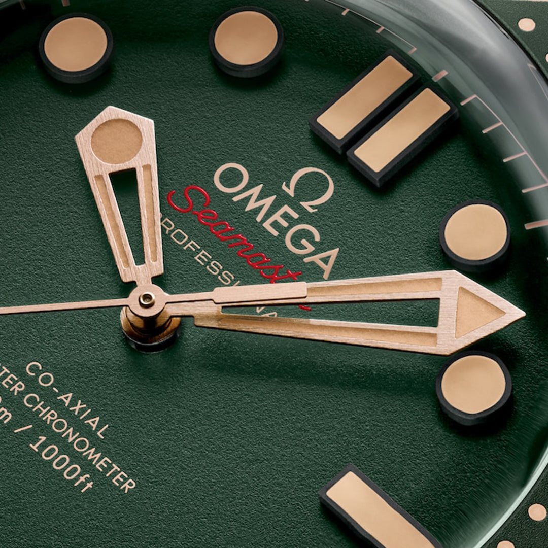 A close-up of the green OMEGA Seamaster Diver 300M Co-Axial Master Chronometer 42mm watch showcases gold hour markers and hands, with "OMEGA Seamaster Diver 300M" in red and white text. Its titanium and Bronze Gold accents add elegance.