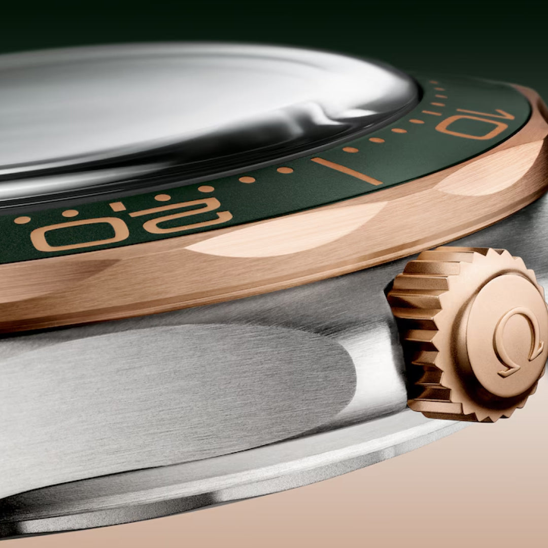 A close-up of the OMEGA Seamaster Diver 300M Co-Axial Master Chronometer 42mm reveals its brushed titanium and Bronze Gold case, green and bronze bezel with numerals, and a prominent gold crown embossed with the logo. The slightly domed crystal gently reflects light on this luxury watch.