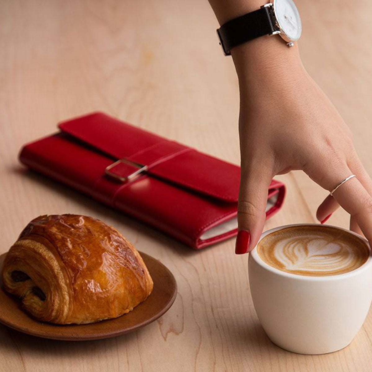 A woman's hand, decorated with a watch and red-painted nails, stretches toward a cup of latte featuring heart-shaped art. Nearby on the wooden table rests a chocolate croissant on a wooden plate next to a red leather wallet and her WOLF 1834 Palermo Jewellery Roll by WOLF, which comes equipped with LusterLoc anti-tarnish technology.