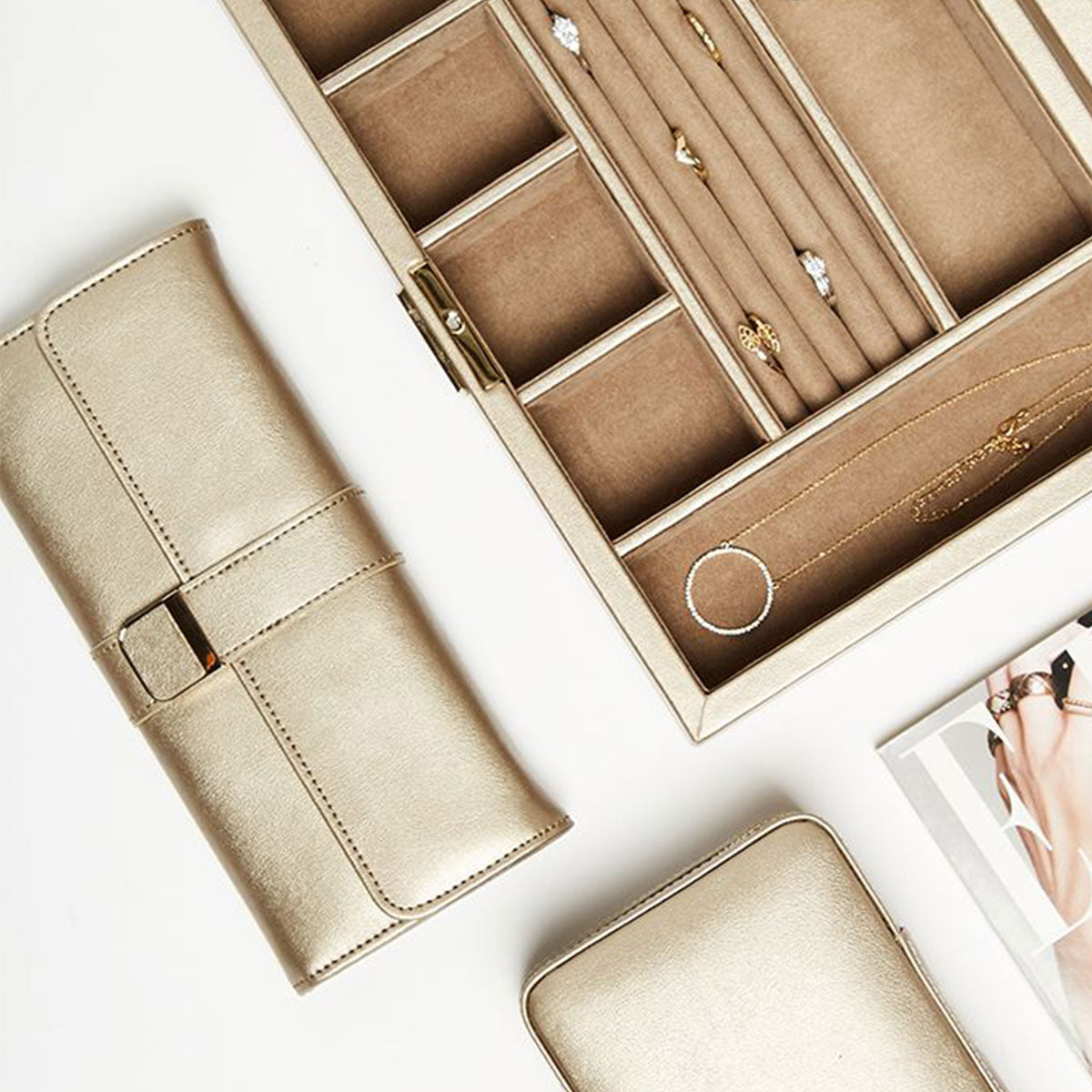 A WOLF 1834 Palermo Jewellery Roll in beige, designed to store necklaces and rings with its anti-tarnish lining to maintain their condition, is open on a flat surface. Nearby, there is a matching travel storage pouch along with a small zippered case. A partial view of a catalog page displaying jewelry images can also be seen.