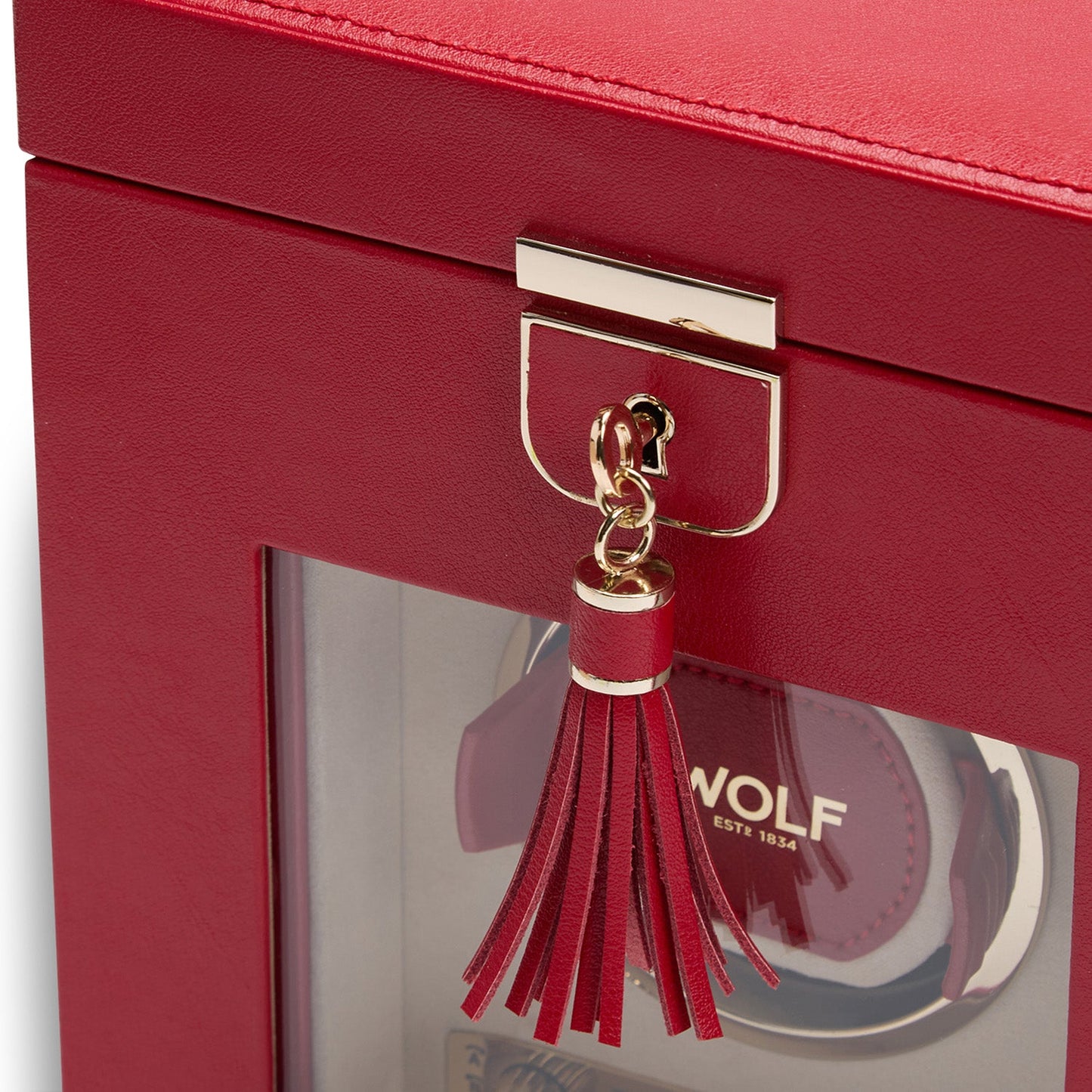 A close-up of the WOLF 1834 Palermo Single Watch Winder with Jewelry Storage in red leather beautifully highlights its sophistication. Its gold clasp and decorative tassel add elegance, while a transparent window reveals the contents inside. Among them is a wristwatch adorned with the visible WOLF brand logo, resting on an integrated watch winder that offers dynamic rotation options.