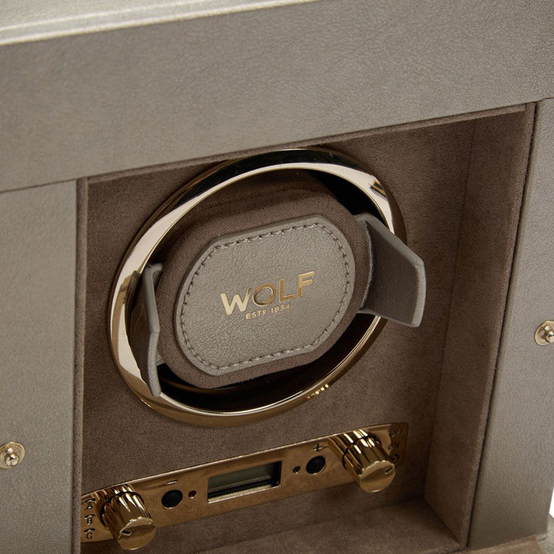 A close-up of the WOLF 1834 Palermo Single Watch Winder with Jewelry Storage reveals a gray cushion inside, adorned with a gold logo that reads "WOLF EST. 1834." This patented innovation boasts metallic controls and rotation options displayed on a small panel, all elegantly positioned within a suede-lined compartment.