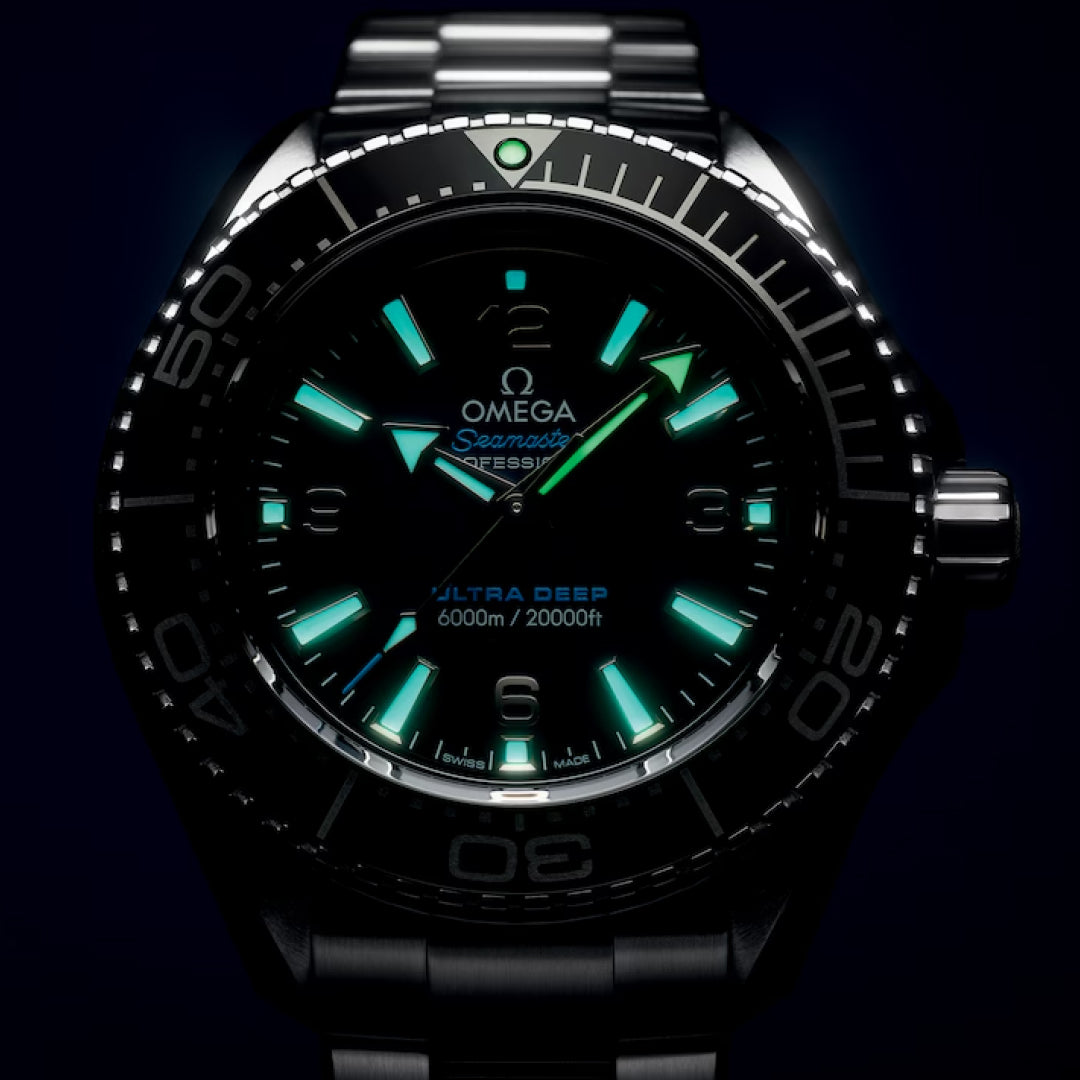 A close-up of the OMEGA Seamaster Planet Ocean 6000M Ultra Deep Co-Axial Master Chronometer 45.5mm shows its O-MEGASTEEL case, metal band, blue and green luminescent markers and hands, and "Ultra Deep 6000m/20000ft" text on the black dial.