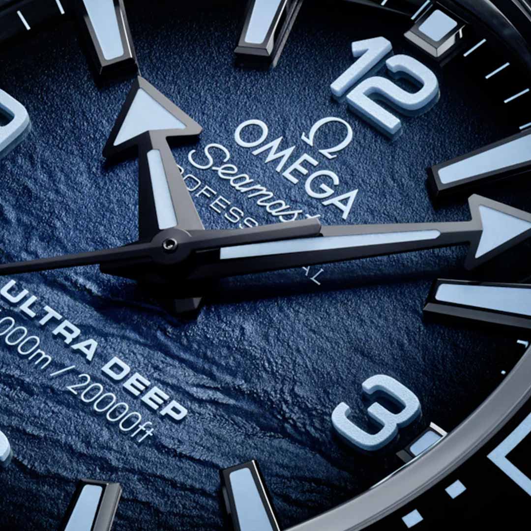 Close-up of an OMEGA Seamaster Planet Ocean 6000M Ultra Deep Co-Axial Master Chronometer 45.5mm watch face, crafted from O-MEGASTEEL, featuring a dark blue textured surface with white hour markers. The hands indicate 6:30. The words "Ultra Deep" and the depth ratings "6000m/20000ft" underscore its distinction as a premier water-resistant timepiece from OMEGA.