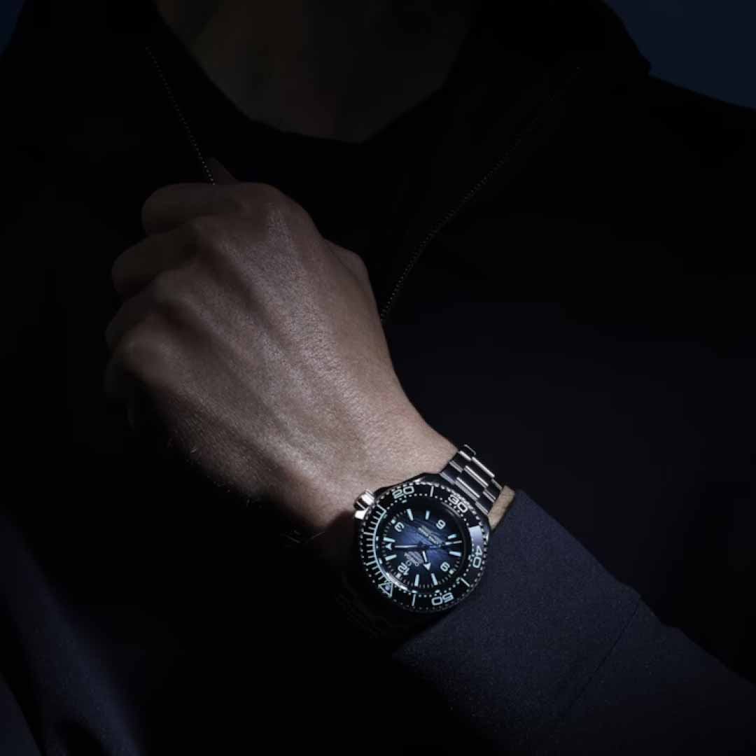 In a dimly lit setting, a person's wrist is adorned with the OMEGA Seamaster Planet Ocean 6000M Ultra Deep Co-Axial Master Chronometer 45.5mm Watch, crafted from O-MEGASTEEL. The watch boasts a black face, luminous hands, and white numerals, with the dark background highlighting this exceptional water-resistant timepiece.