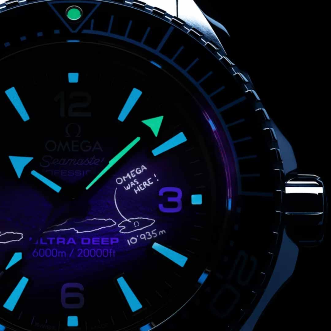 A close-up of the OMEGA Seamaster Planet Ocean 6000M Ultra Deep Co-Axial Master Chronometer 45.5mm watch glowing in the dark. Made from O-MEGASTEEL, it boasts luminescent hands and markers with a drawing and text reading "Omega was here!" on the watch face, exemplifying its status as an outstanding water-resistant timepiece.