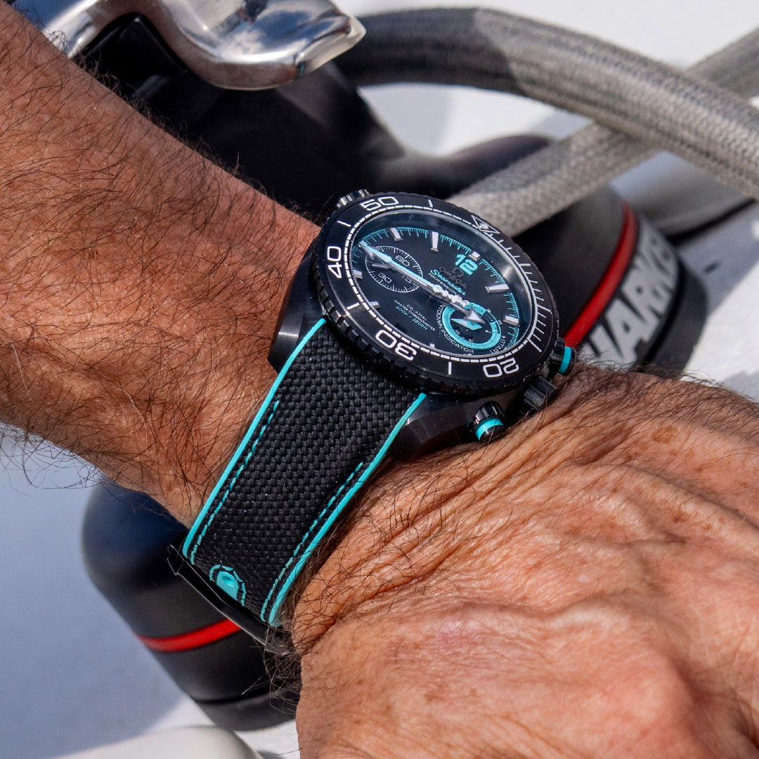 A close-up of a person's wrist showcases the sporty OMEGA Seamaster Planet Ocean 600M Co-Axial Master Chronometer Chronograph 45.5mm Watch, featuring a black ceramic case and teal strap. The watch face is detailed with time markers, a small subdial, and a countdown indicator. Nautical equipment in the background suggests an Emirates Team New Zealand maritime adventure from OMEGA.