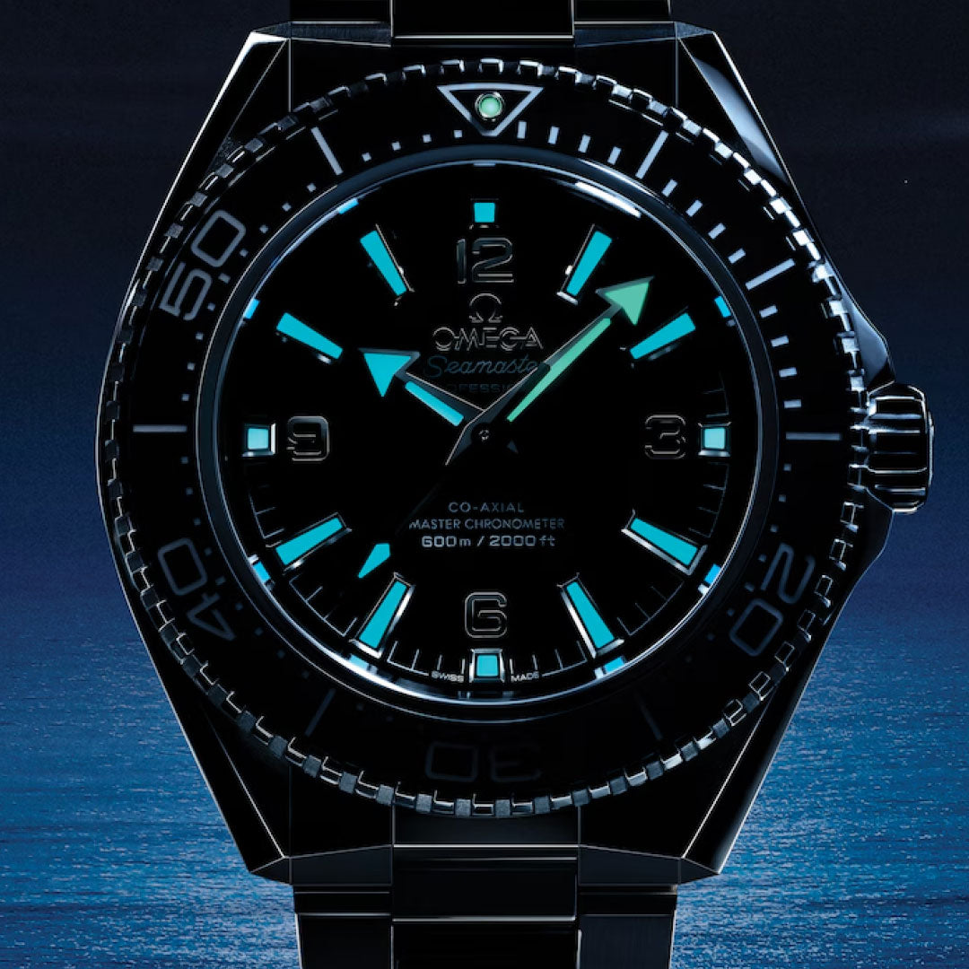 Close-up of the OMEGA Seamaster Planet Ocean 600M Co-Axial Master Chronometer 42mm Watch, featuring a black bezel and luminescent blue and green hands and markers, set against a dark ocean-inspired background.