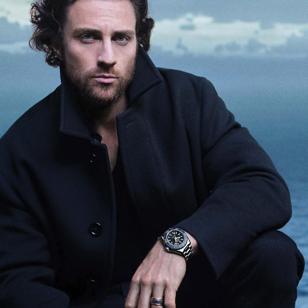 A man with curly brown hair and a trimmed beard, in a dark coat, sits by the sea under cloudy blue skies. He looks at the camera, resting his arm on his knee to display an OMEGA Seamaster Planet Ocean 600M Co-Axial Master Chronometer 42mm Watch.