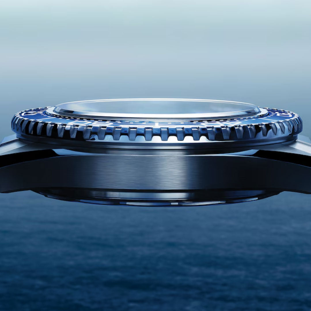 A close-up side view of the OMEGA Seamaster Planet Ocean 600M Co-Axial Master Chronometer 42mm watch bezel, featuring metallic and blue details, set against a blurred ocean and sky backdrop.