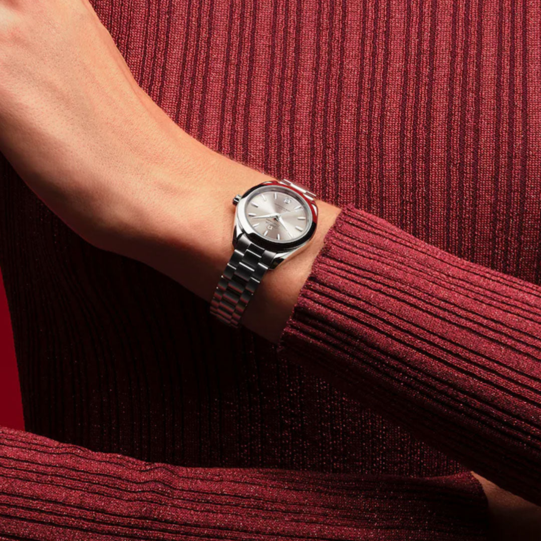 An individual in a ribbed, long-sleeve, dark red top crosses their arm over their chest, showcasing the OMEGA Seamaster Aqua Terra 150M Co-Axial Master Chronometer 30mm Watch with a round white dial.