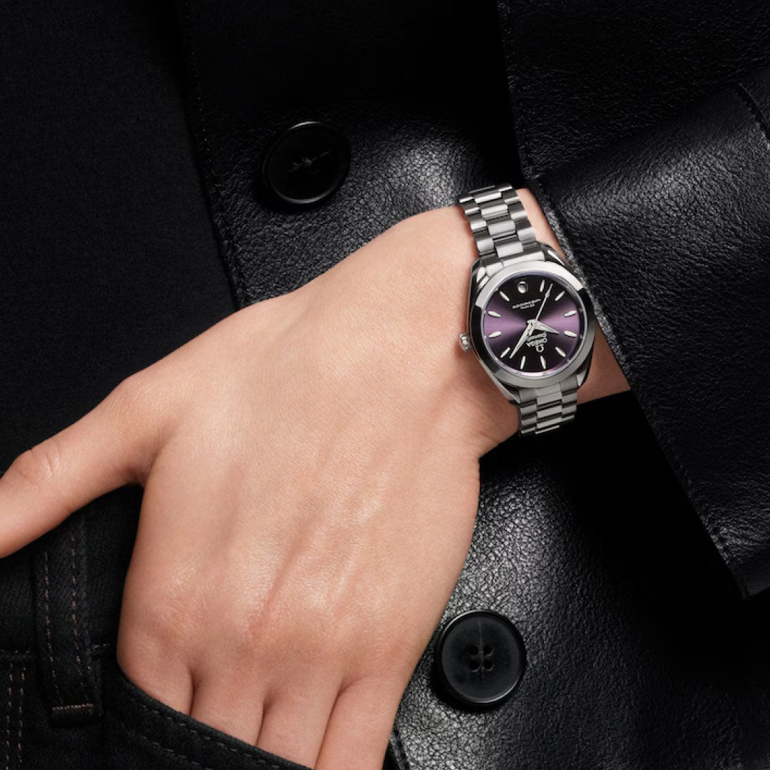 Someone in a black leather jacket and dark pants wears an OMEGA Seamaster Aqua Terra 150M Co-Axial Master Chronometer 30mm watch with a purple dial and metal band on their left wrist, hand relaxed by their hip.
