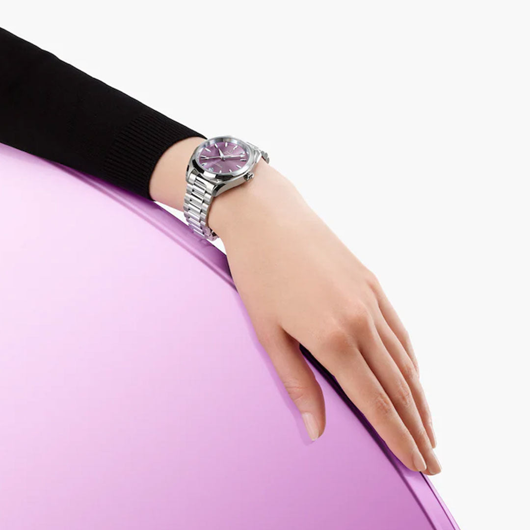 A person’s left hand, in a black long-sleeve top, rests on a shiny lavender surface while wearing the elegant OMEGA Seamaster Aqua Terra Shades Co-Axial Master Chronometer 34mm Watch with a purple dial. The background is white.
