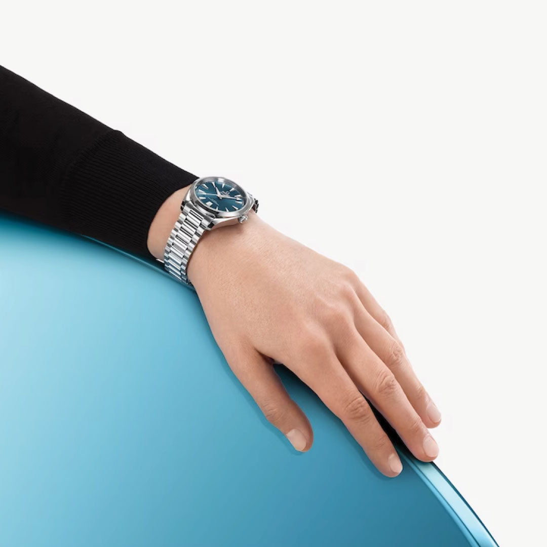 An elegant hand adorned with an OMEGA Seamaster Aqua Terra Shades Co-Axial Master Chronometer 38mm Watch with a blue dial gently places itself on a sleek, curved light blue surface. The timepiece subtly emerges from beneath the black sleeve, contrasting strikingly against the simple white backdrop.