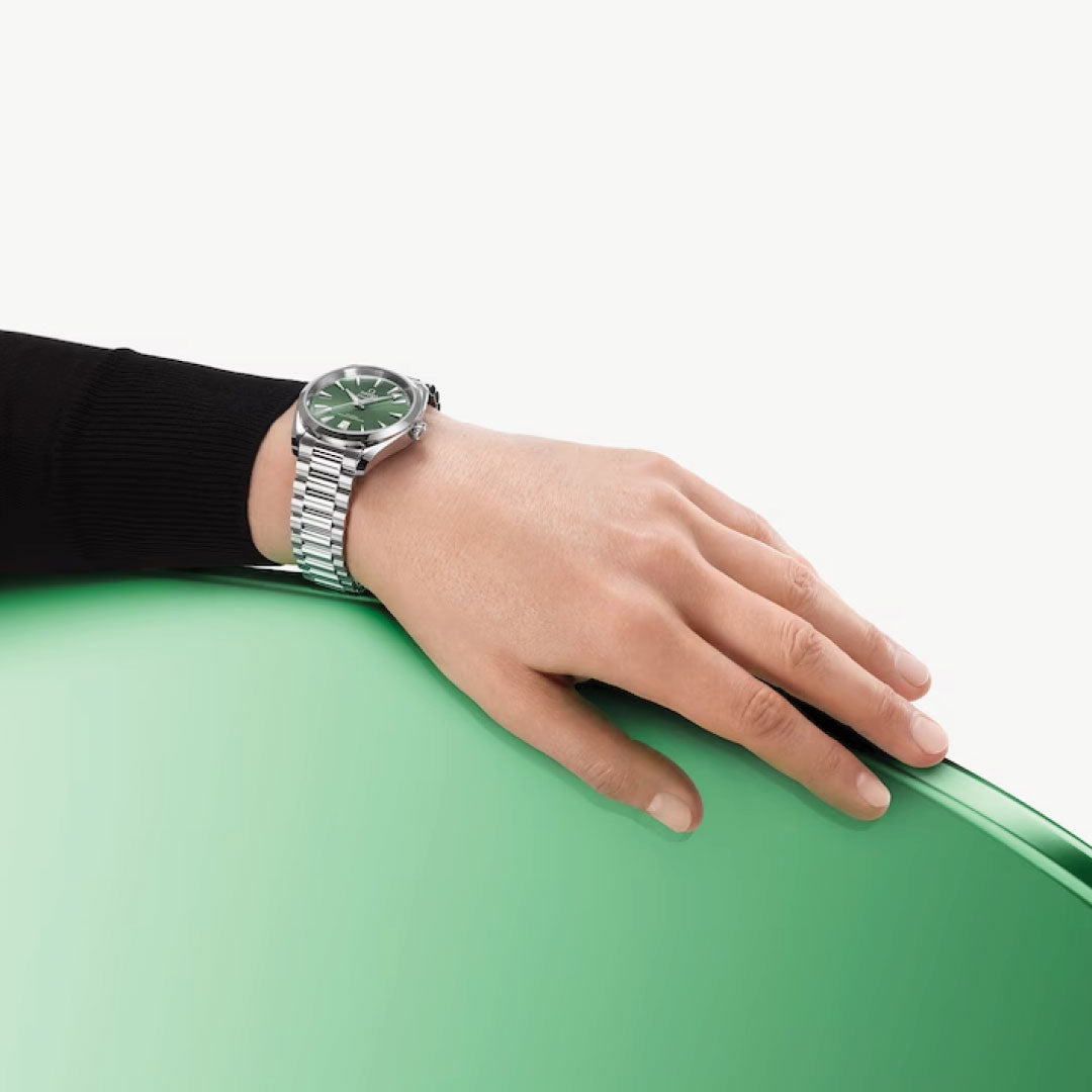 A person wearing an OMEGA Seamaster Aqua Terra Shades Co-Axial Master Chronometer 38mm watch, featuring a green face, rests their hand on a glossy green surface. The background is light beige. The relaxed hand highlights the metallic band of the watch, subtly gleaming and emphasizing its Master Chronometer calibre 8800 precision.