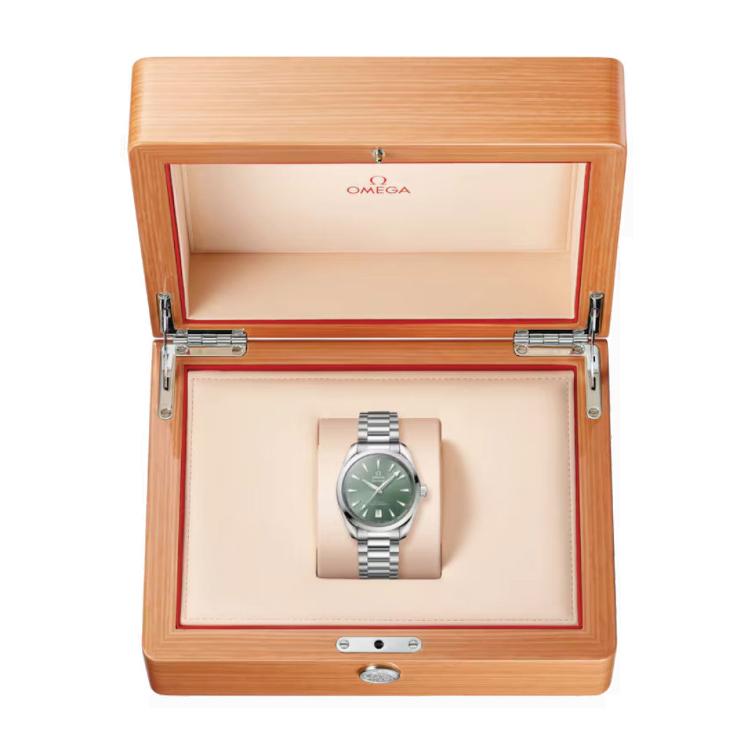 A silver OMEGA Seamaster Aqua Terra Shades Co-Axial Master Chronometer 38mm watch with a green dial is elegantly showcased inside a wooden box with a beige interior. The open lid prominently displays the OMEGA logo, emphasizing the sophistication of this timepiece. It features a metal link band and boasts the Master Chronometer calibre 8800.