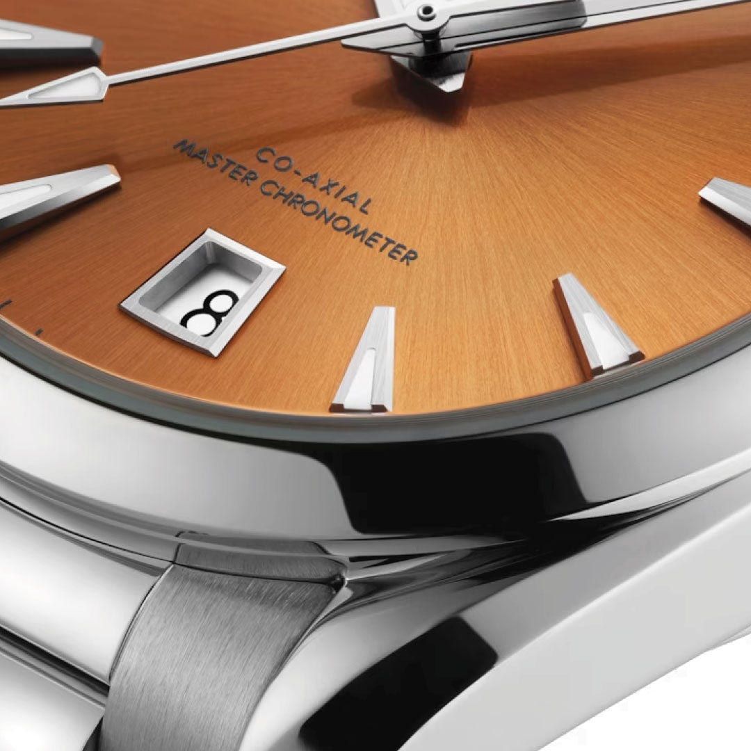 A detailed view of the OMEGA Seamaster Aqua Terra Shades Co-Axial Master Chronometer 38mm watch showcases a luxurious sunburst orange dial, accented with silver hour markers and a date window displaying the number 8. The elegant silver bezel highlights the inscription "Co-Axial Master Chronometer," embodying OMEGA's maritime heritage and the refined elegance of the Seamaster Aqua Terra collection.