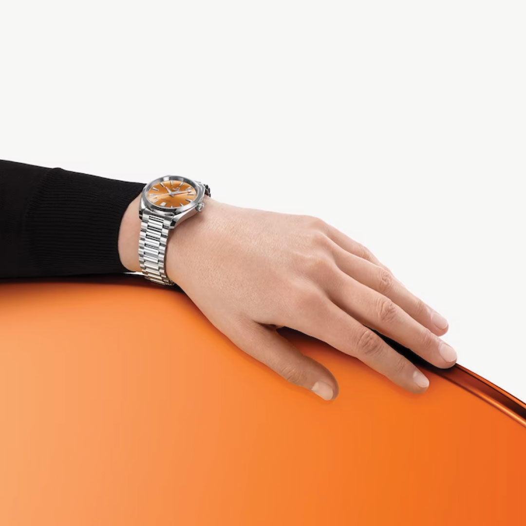 A hand wearing the OMEGA Seamaster Aqua Terra Shades Co-Axial Master Chronometer 38mm Watch, distinguished by its silver case and striking orange dial, rests on a vivid orange surface against a simple, light backdrop. The black sleeve of the wearer enhances the watch's dynamic design, highlighting its maritime roots and precision as part of OMEGA's esteemed collection.