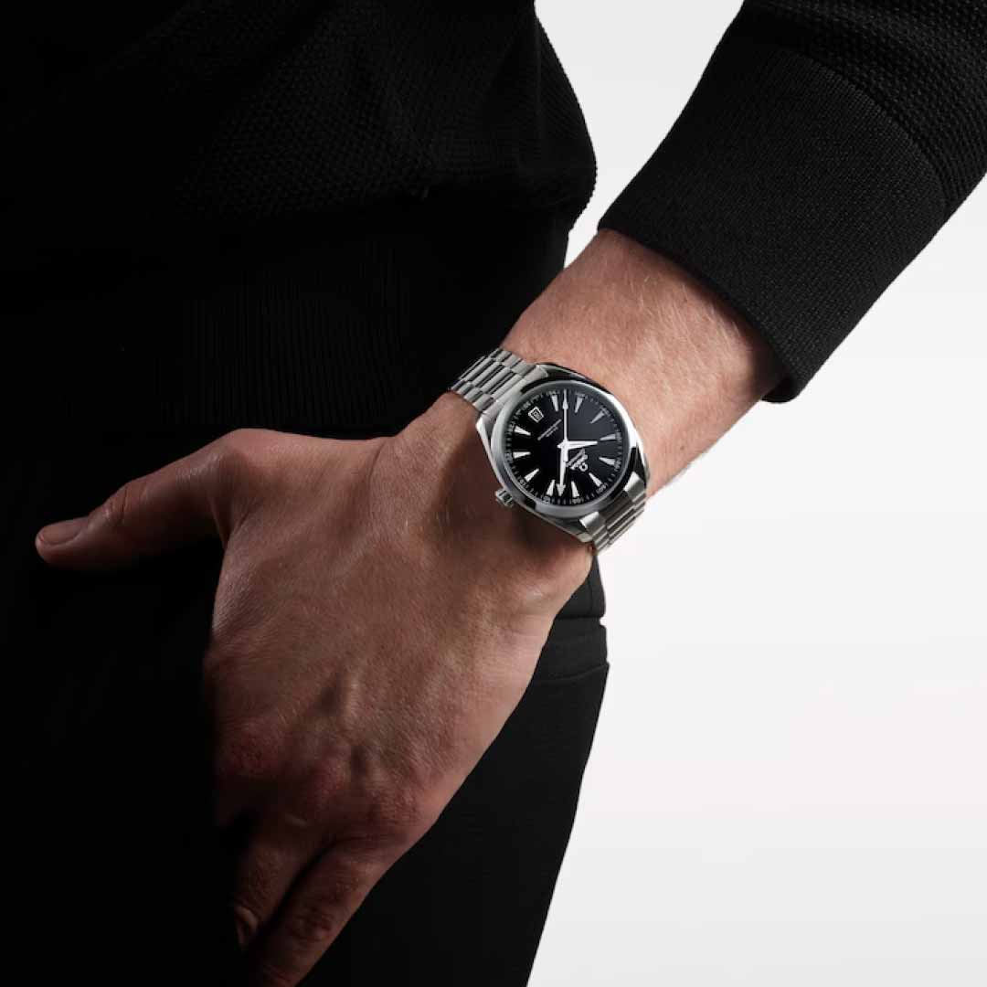 An individual is sporting a stylish OMEGA Seamaster Aqua Terra 150m Co-Axial Master Chronometer 41mm watch, featuring a black dial adorned with silver hour markers. Their hand is relaxed, resting by their side against a white and gray backdrop while wearing a dark sweater.