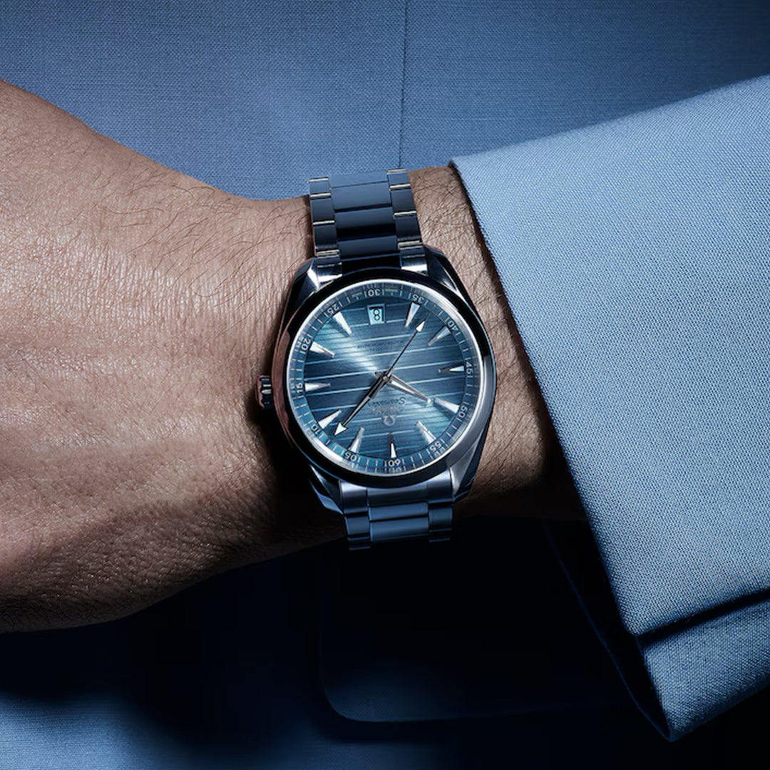 A close-up of a wrist wearing the OMEGA Seamaster Aqua Terra 150M Co-Axial Master Chronometer 41mm with a sunburst dial and date display. The blue metallic watch features a metal strap against a blue long-sleeve shirt, its reflective surface highlighted by the lighting.