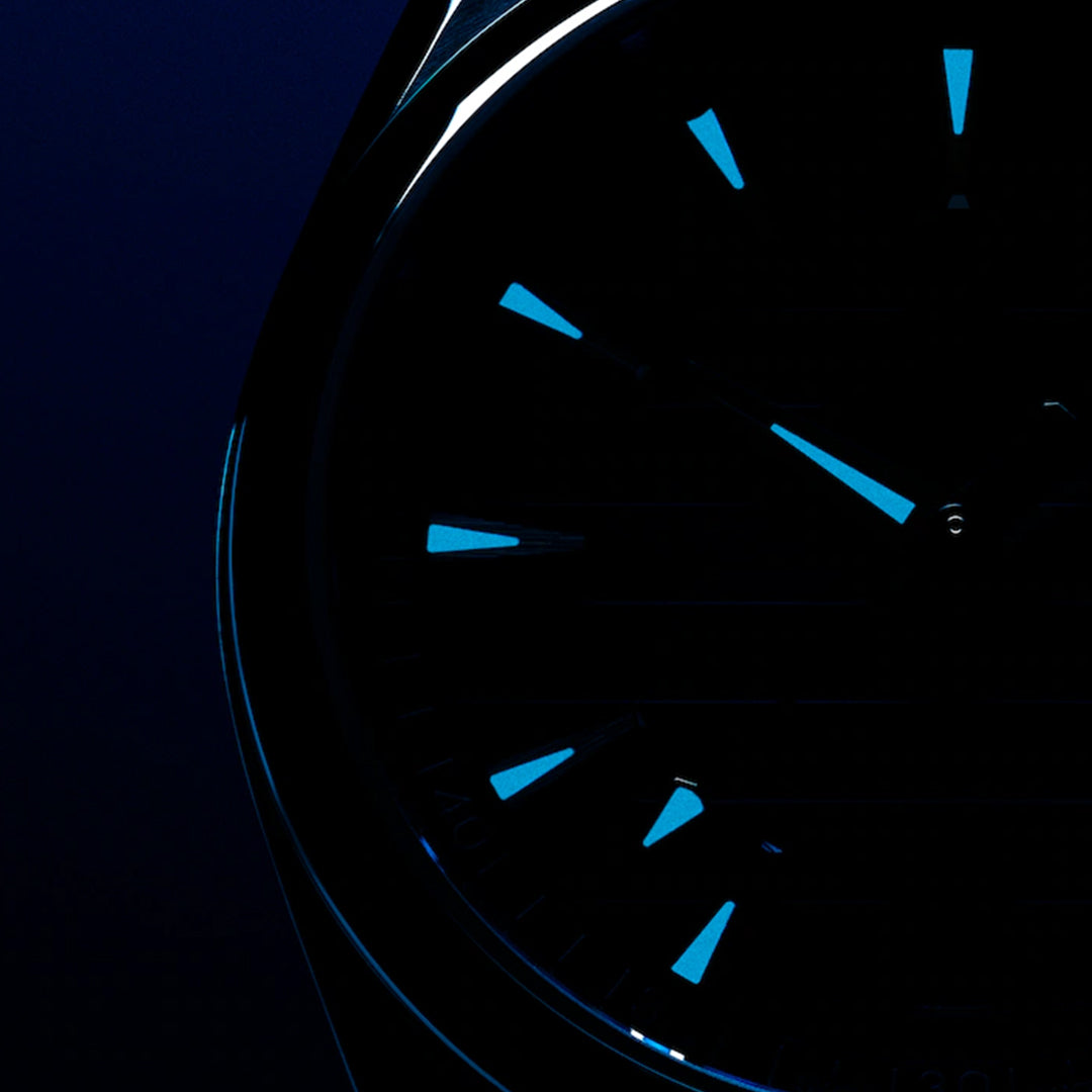 Close-up of an OMEGA Seamaster Aqua Terra 150M Co-Axial Master Chronometer 41mm watch face in dim lighting, showcasing luminescent blue hour markers and hands against a dark background, with the left side of the case faintly visible.