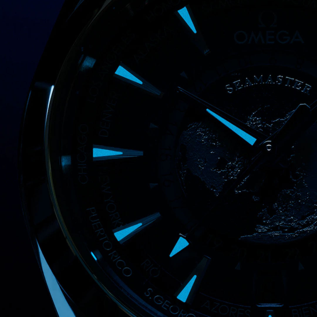 Close-up of the OMEGA Seamaster Aqua Terra 150M Worldtimer Co-Axial Master Chronometer 43mm Summer Blue Edition watch face in low light, highlighting blue luminescent markers, faint city names around the edge, and continents on the dial.