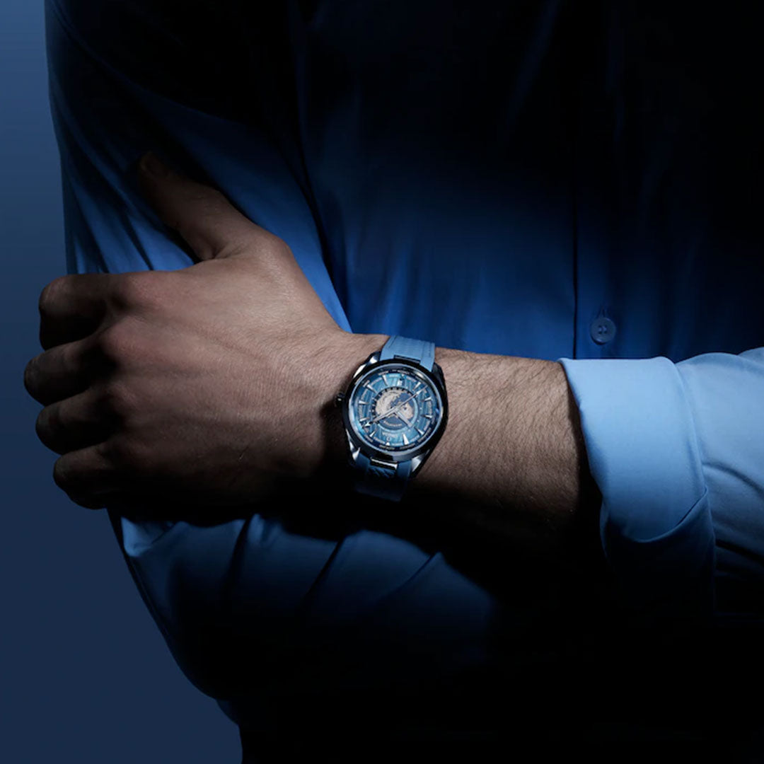 A person with crossed arms wears a blue dress shirt and the OMEGA Seamaster Aqua Terra 150M Worldtimer Co-Axial Master Chronometer 43mm Summer Blue Edition, highlighted under dramatic lighting against a dark background.