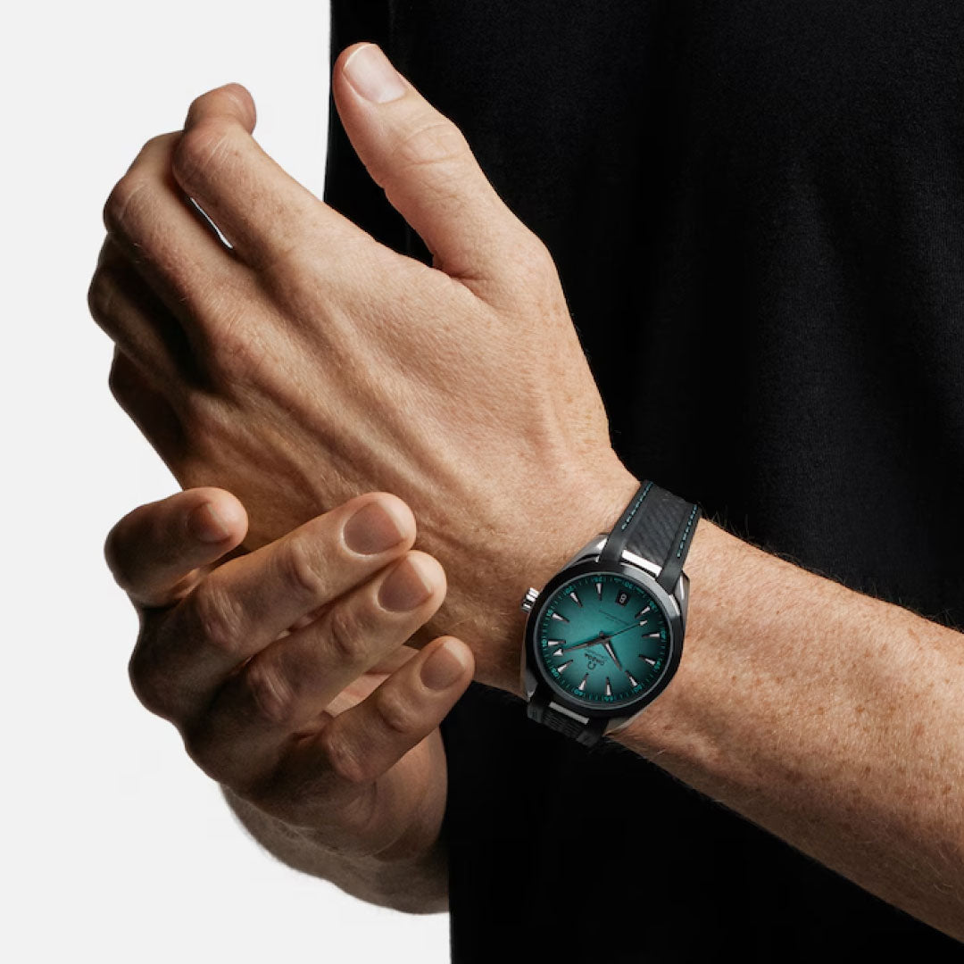 A person in a black shirt showcases an OMEGA Seamaster Aqua Terra 150M Co-Axial Master Chronometer 41mm watch with a green face and black strap on their left wrist, gently touching it with their right hand.