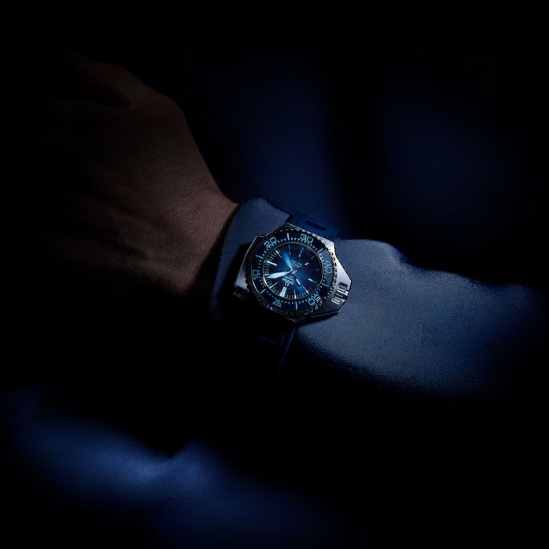 A close-up of a wrist wearing the OMEGA Seamaster Ploprof 1200M Co-Axial Master Chronometer 55mm x 45mm, featuring its illuminated Swiss Made dial and black strap in low light against a dark background.