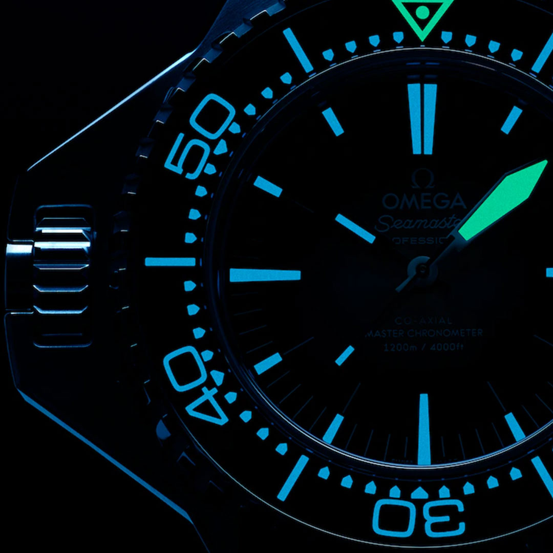 Close-up of the OMEGA Seamaster Ploprof 1200M Co-Axial Master Chronometer watch face glowing blue in the dark, featuring luminescent hour markers and hands, partial O-MEGASTEEL bezel and crown, and “OMEGA Seamaster” text.