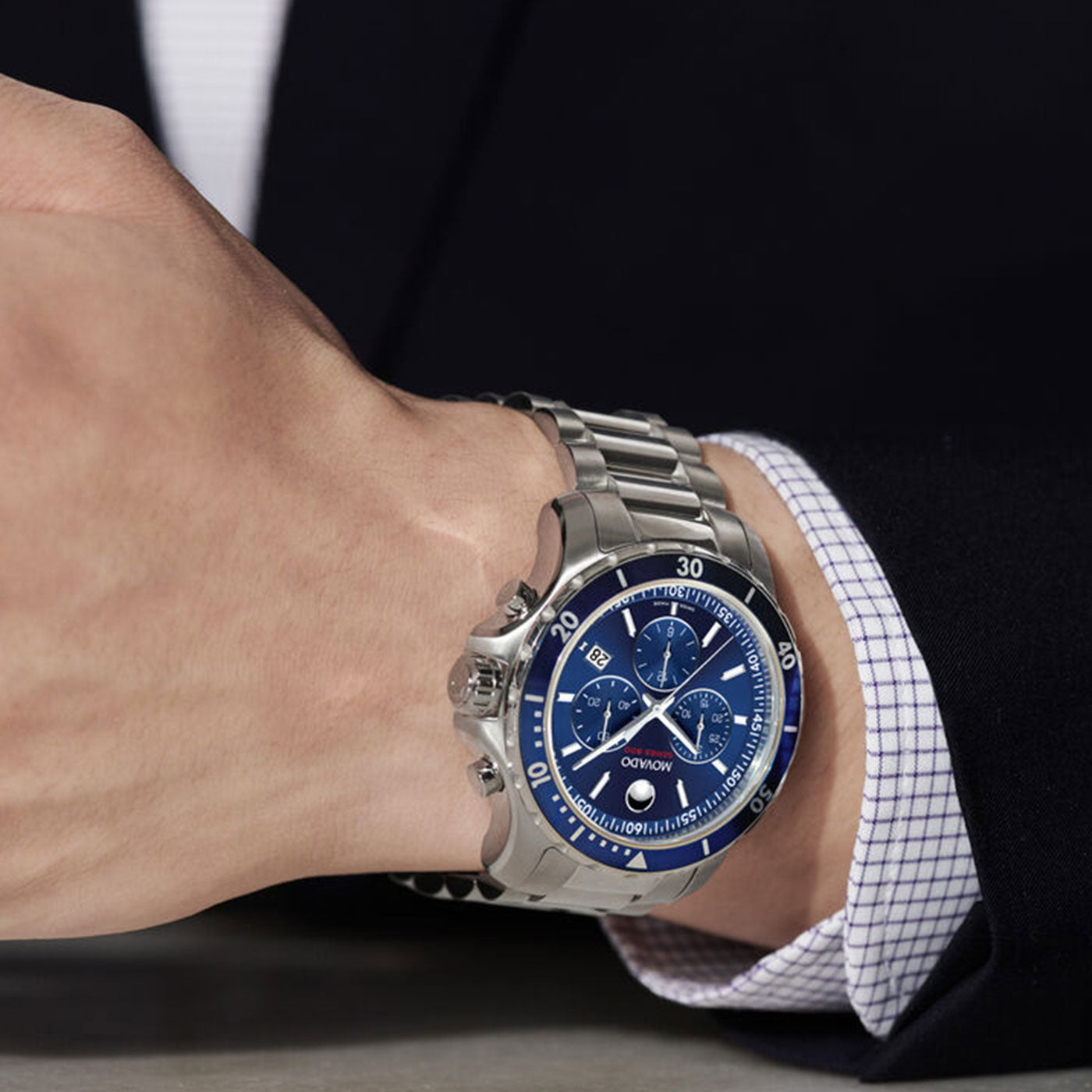 A person wearing a formal suit and a silver wristwatch with a blue chronograph dial featuring multiple subdials. The Movado Series 800 Quartz 40mm Watch, crafted by Movado in Performance Steel, includes a metal bracelet and is stylishly worn on the left wrist.