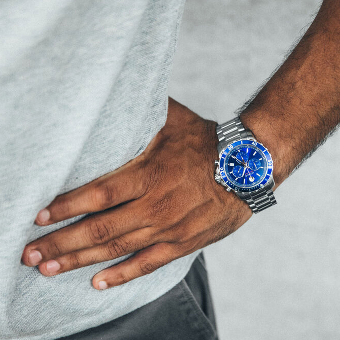 A person wearing a Movado Series 800 Quartz 40mm Watch, featuring a sleek silver stainless steel band and an eye-catching blue chronograph dial, rests their hand on their hip. They are dressed in a light gray shirt and dark pants, perfectly exhibiting an impeccable blend of style and performance steel elegance.