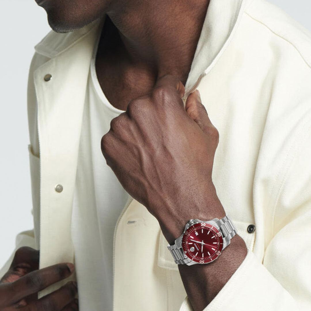 A person in a cream jacket and white shirt showcases the Movado Series 800 Quartz 40mm watch, featuring a striking red face and unidirectional rotating bezel, on their left wrist while touching their jacket collar.