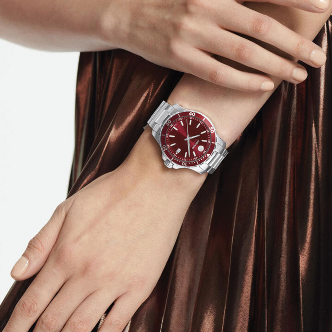 A person wearing the Movado Series 800 Quartz 40mm metallic red and silver watch with a unidirectional rotating bezel rests one hand on the other, dressed in a shiny, brown pleated garment.