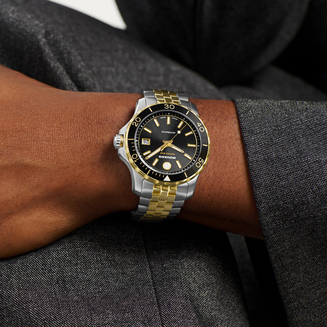 A person dressed in a dark suit is wearing the Movado Series 800 Automatic 40mm Watch by Movado, which features Swiss-made automatic movement, a two-tone stainless steel case, a silver and gold wristband, a black dial with gold accents, and a date display.