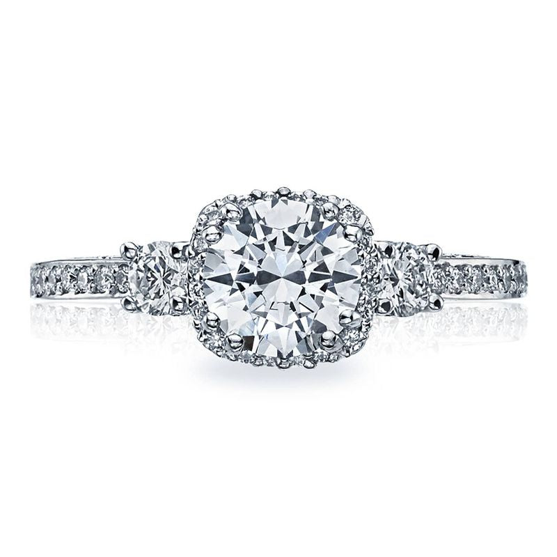 Introducing the Tacori Dantela engagement ring, a stunning piece crafted from 18K white gold. This exquisite three-stone semi-mount design highlights a brilliant round center stone encircled by a halo of smaller diamonds. The elegant band is adorned with additional diamonds, enhancing its overall brilliance and sophistication.