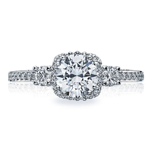 Introducing the Tacori Dantela engagement ring, a stunning piece crafted from 18K white gold. This exquisite three-stone semi-mount design highlights a brilliant round center stone encircled by a halo of smaller diamonds. The elegant band is adorned with additional diamonds, enhancing its overall brilliance and sophistication.