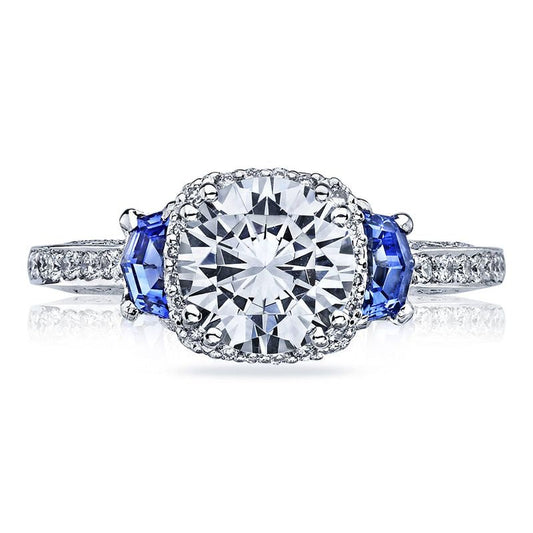 The Tacori Dantela 18K White Gold Round Brilliant Semi Mount Three Stone Hidden Halo Engagement Ring showcases a large central round diamond, flanked by two elegant oval sapphire side stones. The diamond band adds an extra touch of sparkle and sophistication to this stunning design.