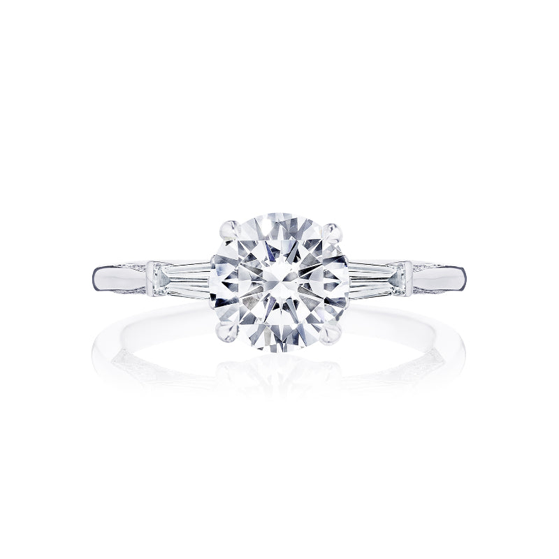 An 18K white gold engagement ring by Tacori, featuring a round brilliant center diamond accented with side stones and a diamond accent band. The elegant design echoes the classic Tacori crescent style, set against a plain white background.